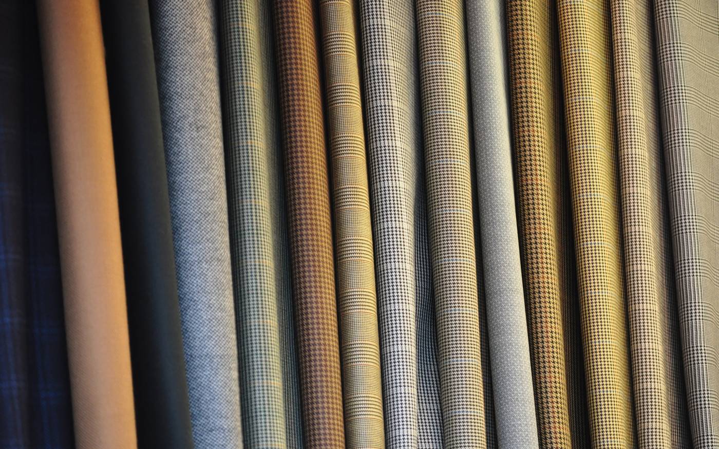 Rotoli di tessuto da abito appesi uno accanto all'altro in verticale: tinte unite cammello, verde scuro e grigio, poi pied de poule e principe di Galles in marrone, senape, grigio e nero
